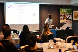 Female presenter in front of audience and backdrop of meeting programme shown on screen