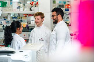 Three students in white lab coats in a lab having a conversation and smiling